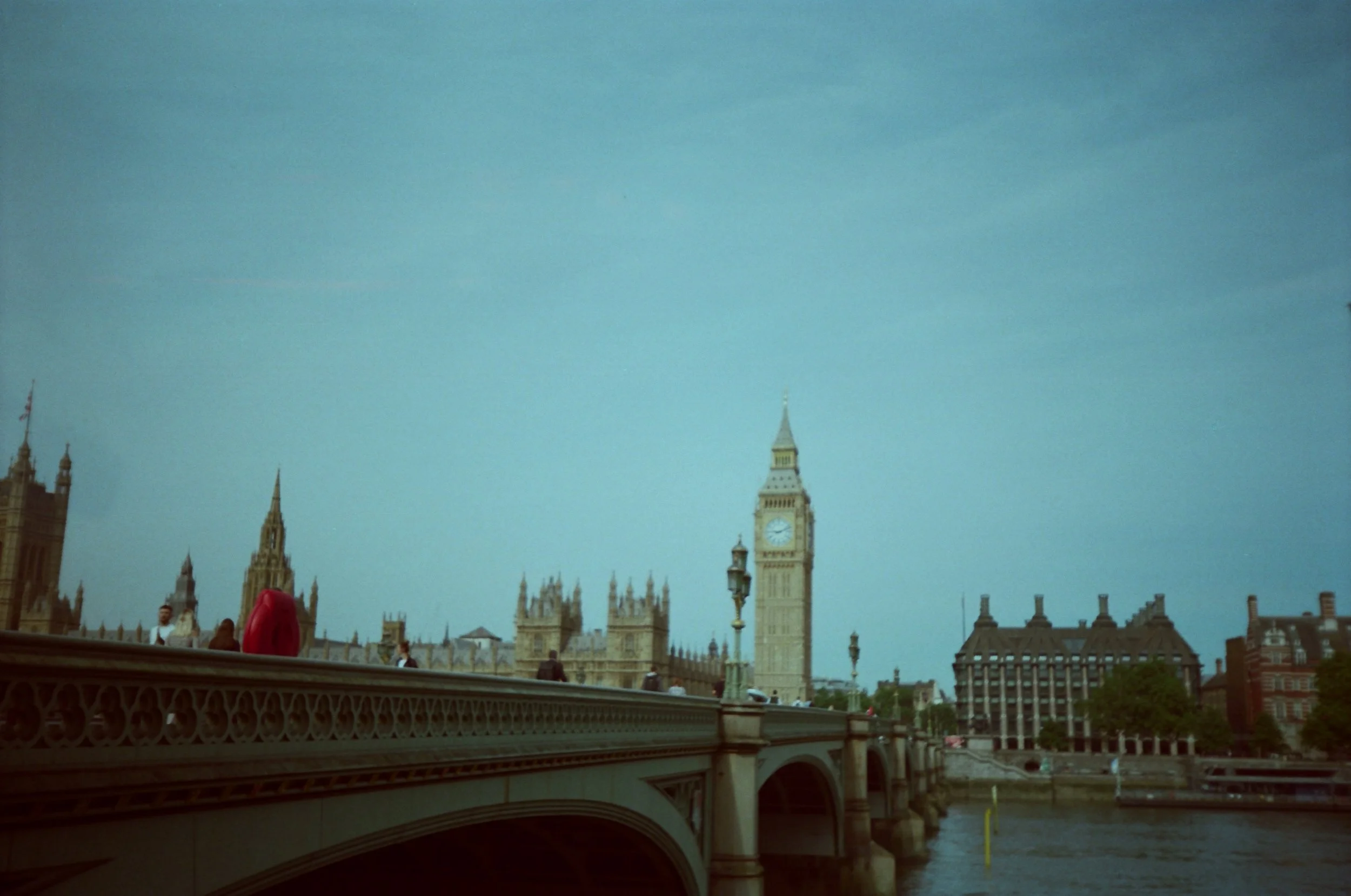 The image shows a view of the Palace of Westminster in London, England, with Big Ben clock tower in the background, over a bridge crossing the River Thames, with a few pedestrians and cloudy sky.