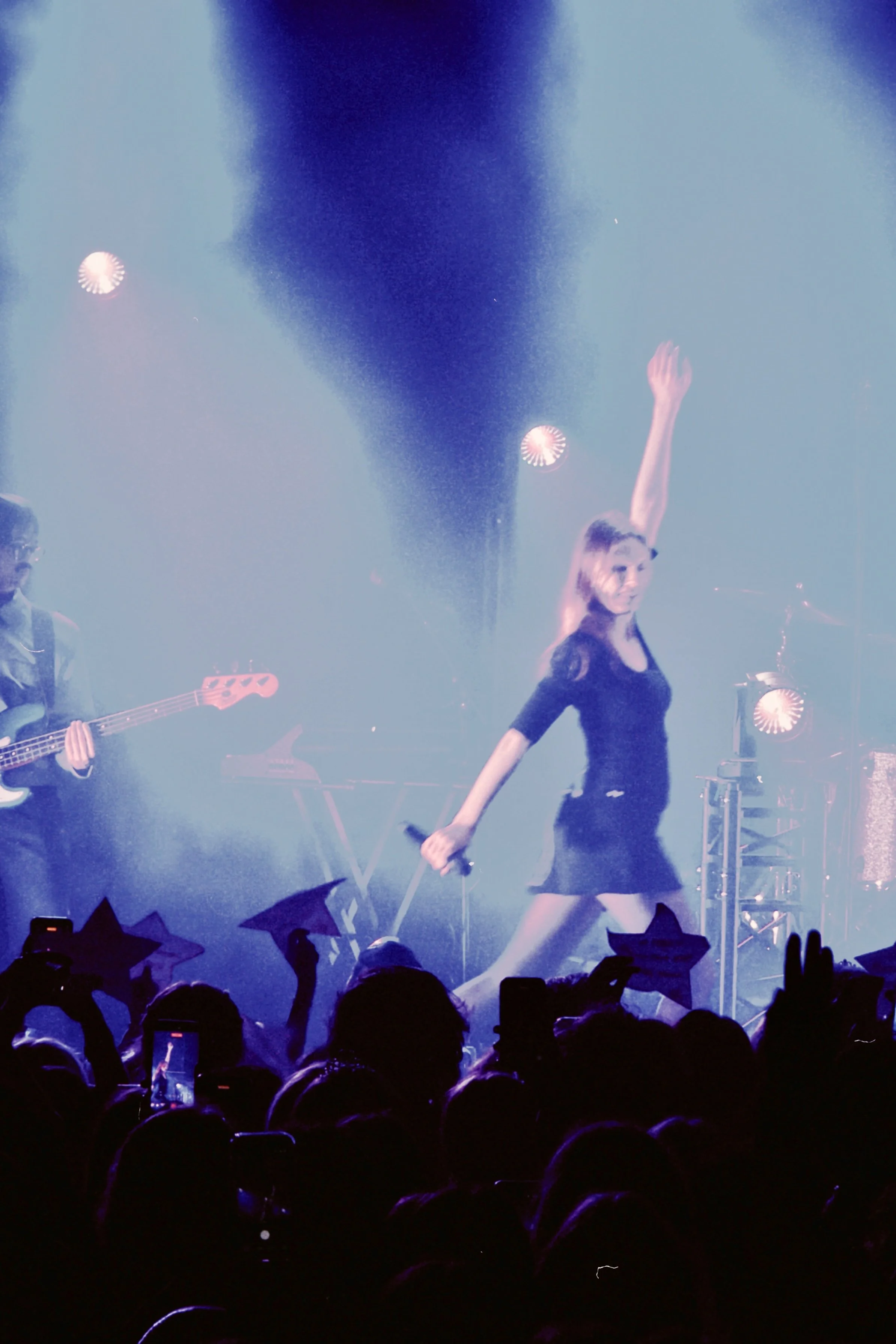 A female singer with blonde hair on stage, dancing with her right arm raised, performing at a concert with colored stage lights and a crowd holding umbrellas.