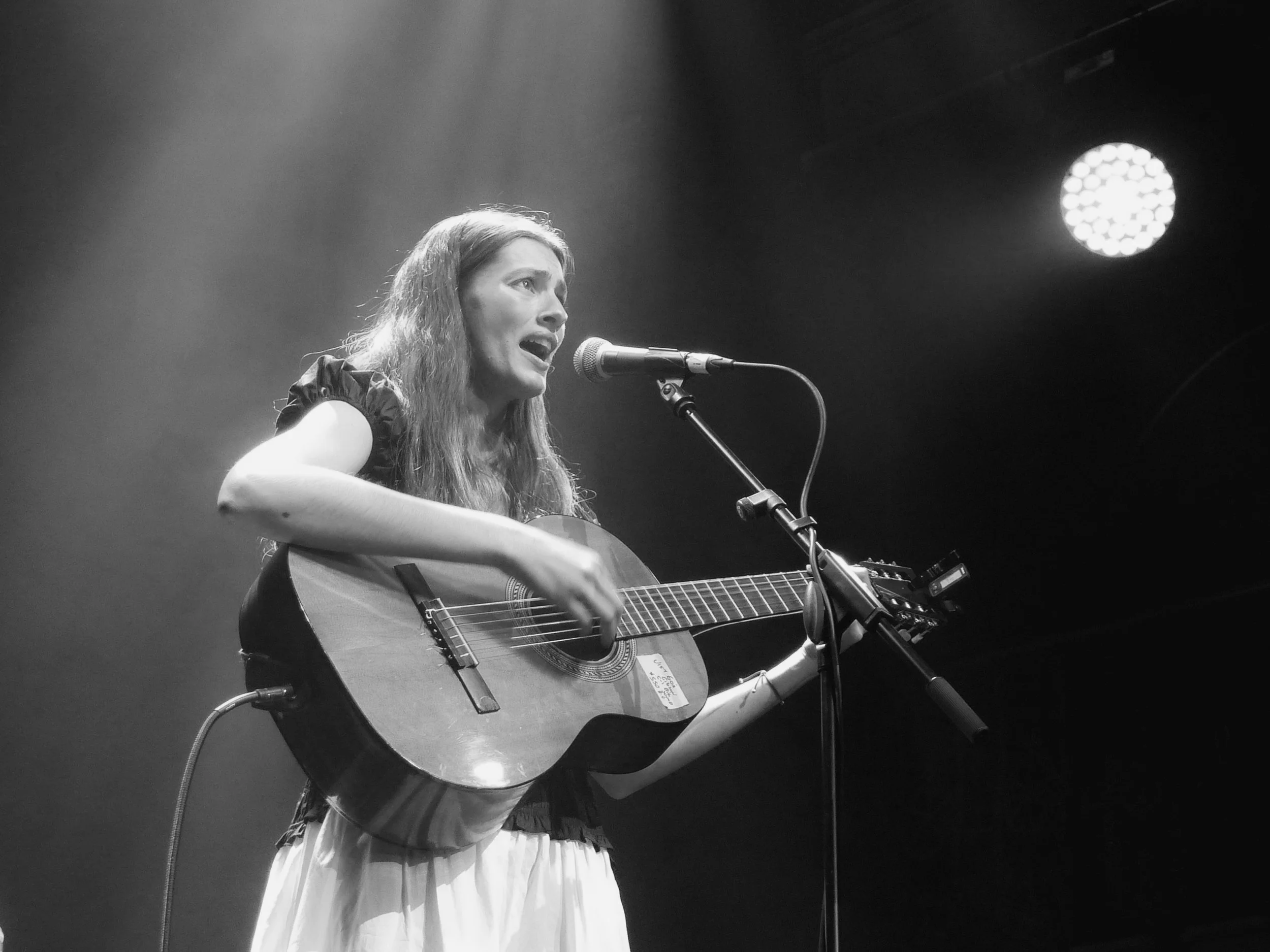 A woman with long hair singing into a microphone while playing an acoustic guitar on stage, with stage lights overhead.