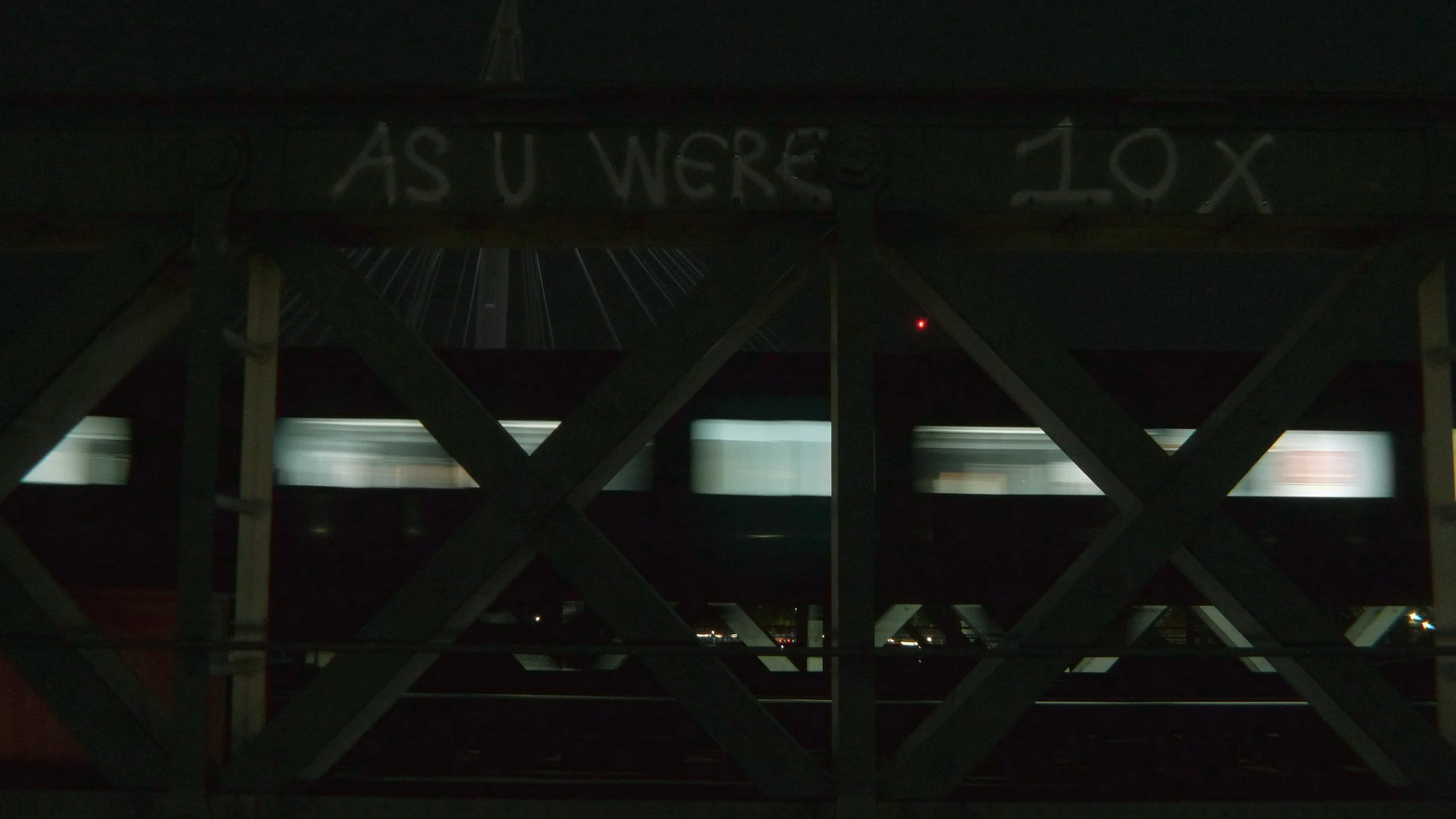 Nighttime view from under a bridge showing a train passing by below, with graffiti on the bridge structure that reads 'AS U WERE 10X' and city lights in the background.