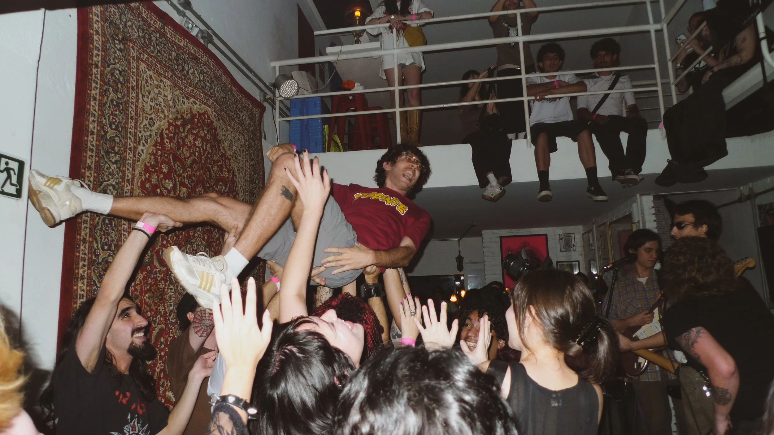 A man with curly hair and glasses, wearing a red shirt, is being lifted by the crowd at a concert or party. People around are reaching out and cheering, and several individuals are sitting on a balcony above observing the scene. The room has a vintag