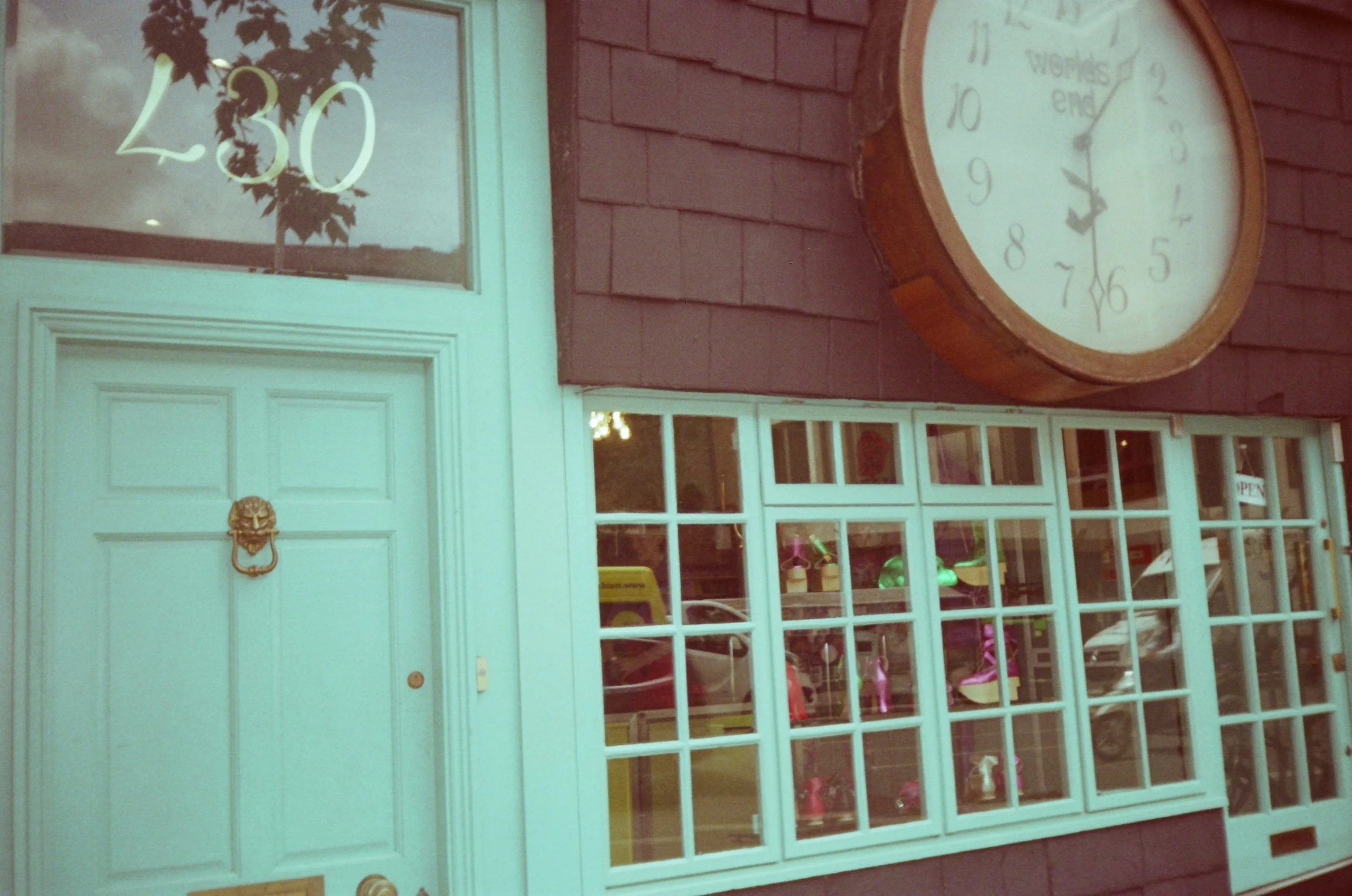Storefront with glass window showing toys and items inside, a white door with gold lion door knocker, and a round clock with roman numerals and a message that reads 'world's end' outside, against a brick wall.
