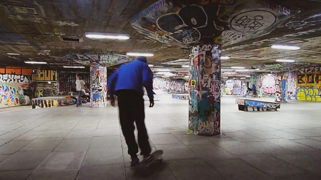 A skateboarder in a blue jacket skateboarding in a graffiti-covered underground tunnel with multiple pillars and benches.