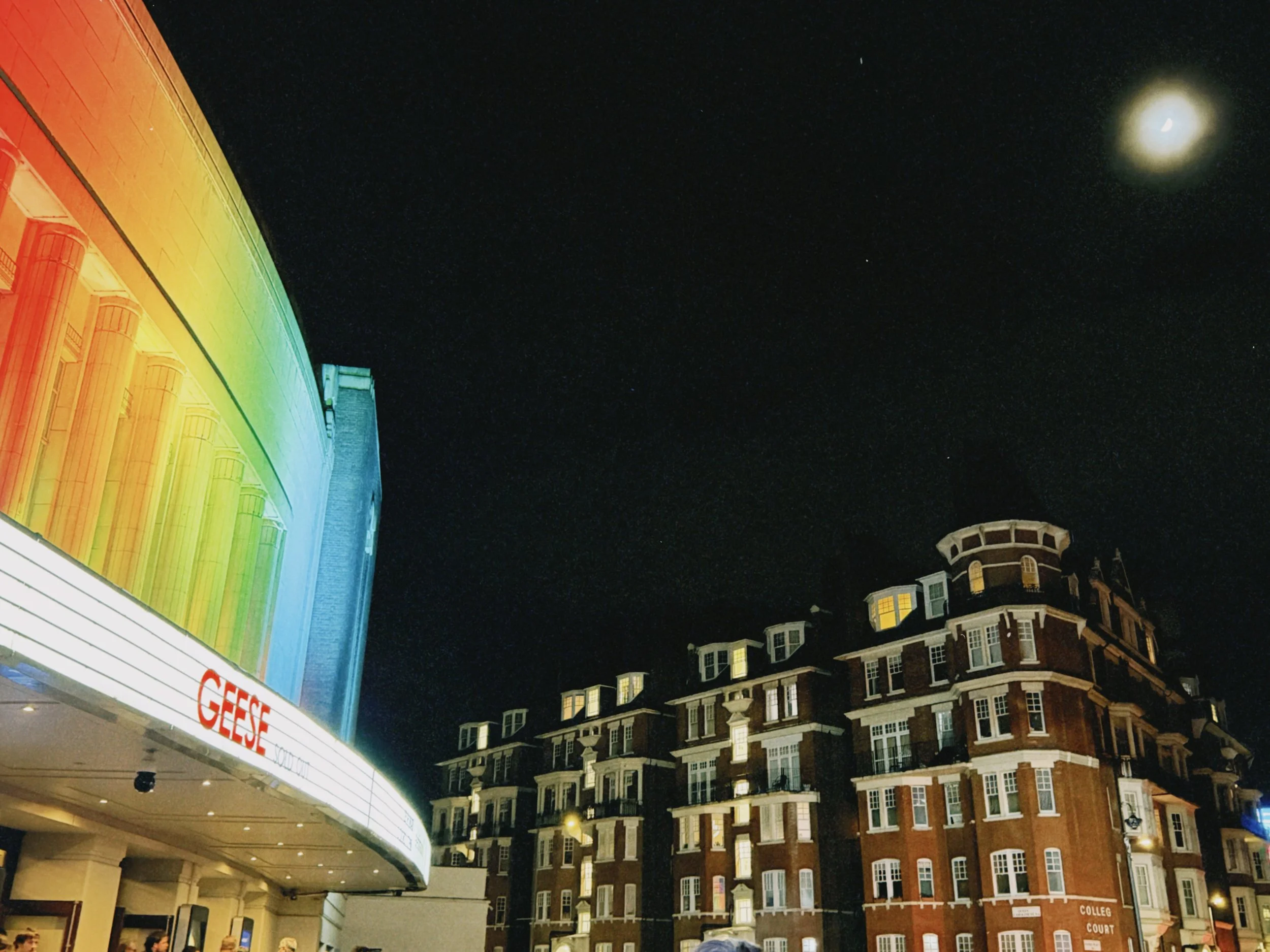 Night view of buildings with Victorian architectural style, illuminated windows, and a large billboard displaying a rainbow-colored image and text that reads 'GEORGE'. The sky is dark with visible stars and the moon.