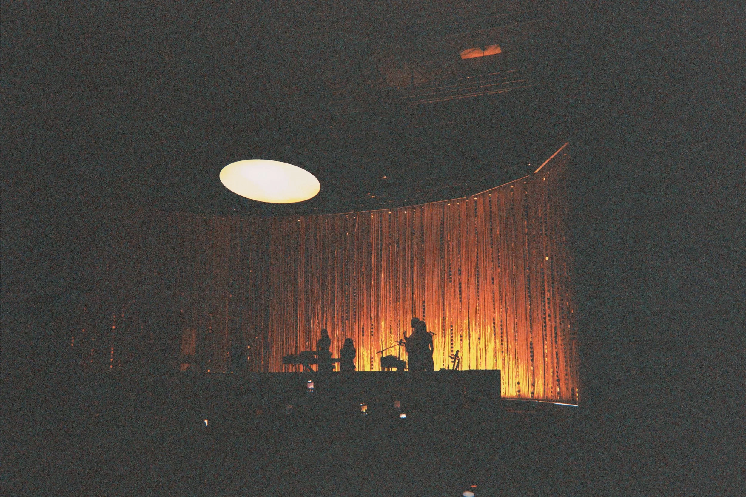 People are performing on stage, silhouetted against a backdrop of curtains illuminated by warm orange lighting. The scene is dark with a large, round, white ceiling light visible.