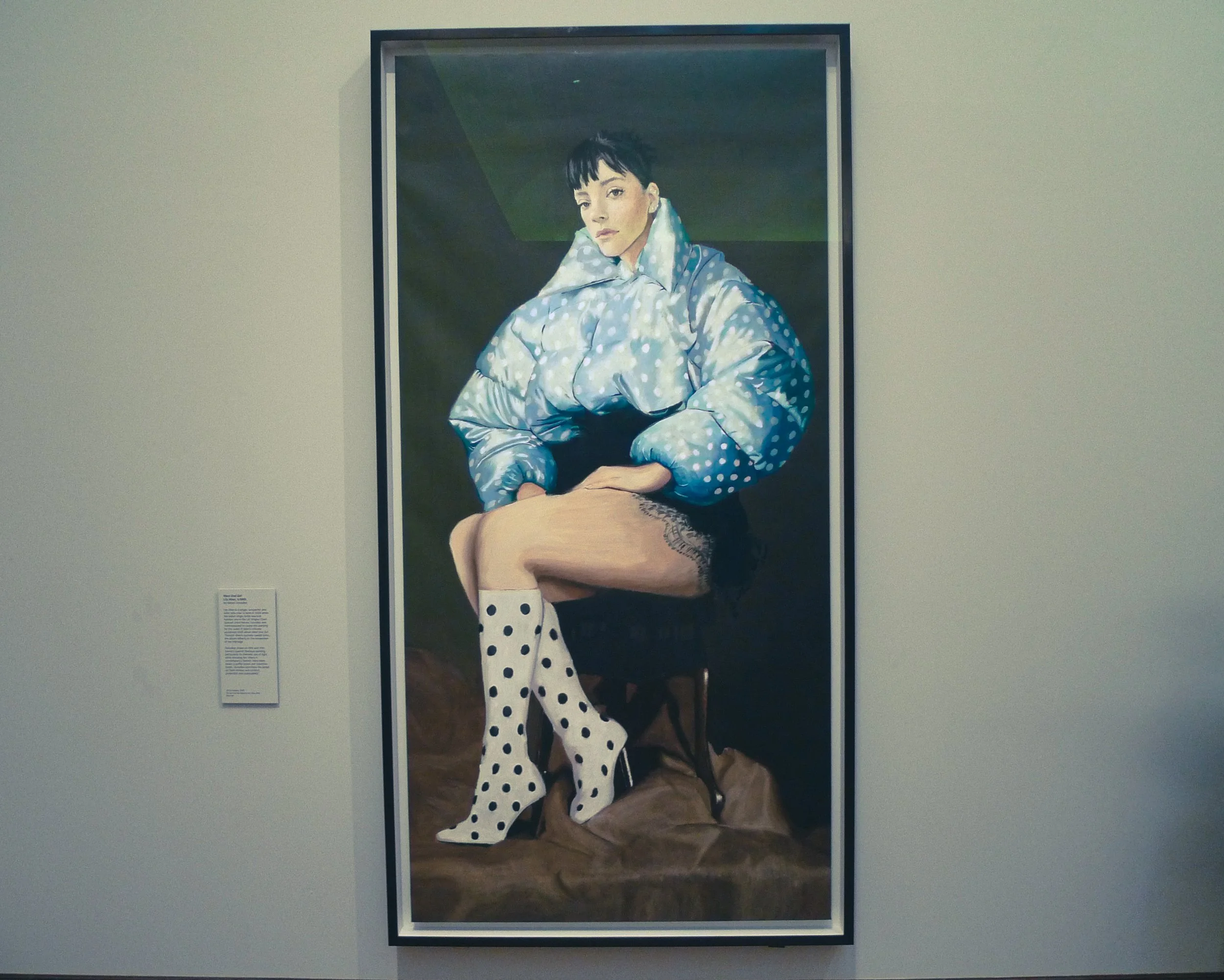 A portrait painting of a woman with black hair and bangs, wearing a blue polka dot coat and matching polka dot stockings, sitting on a wooden surface against a dark background.