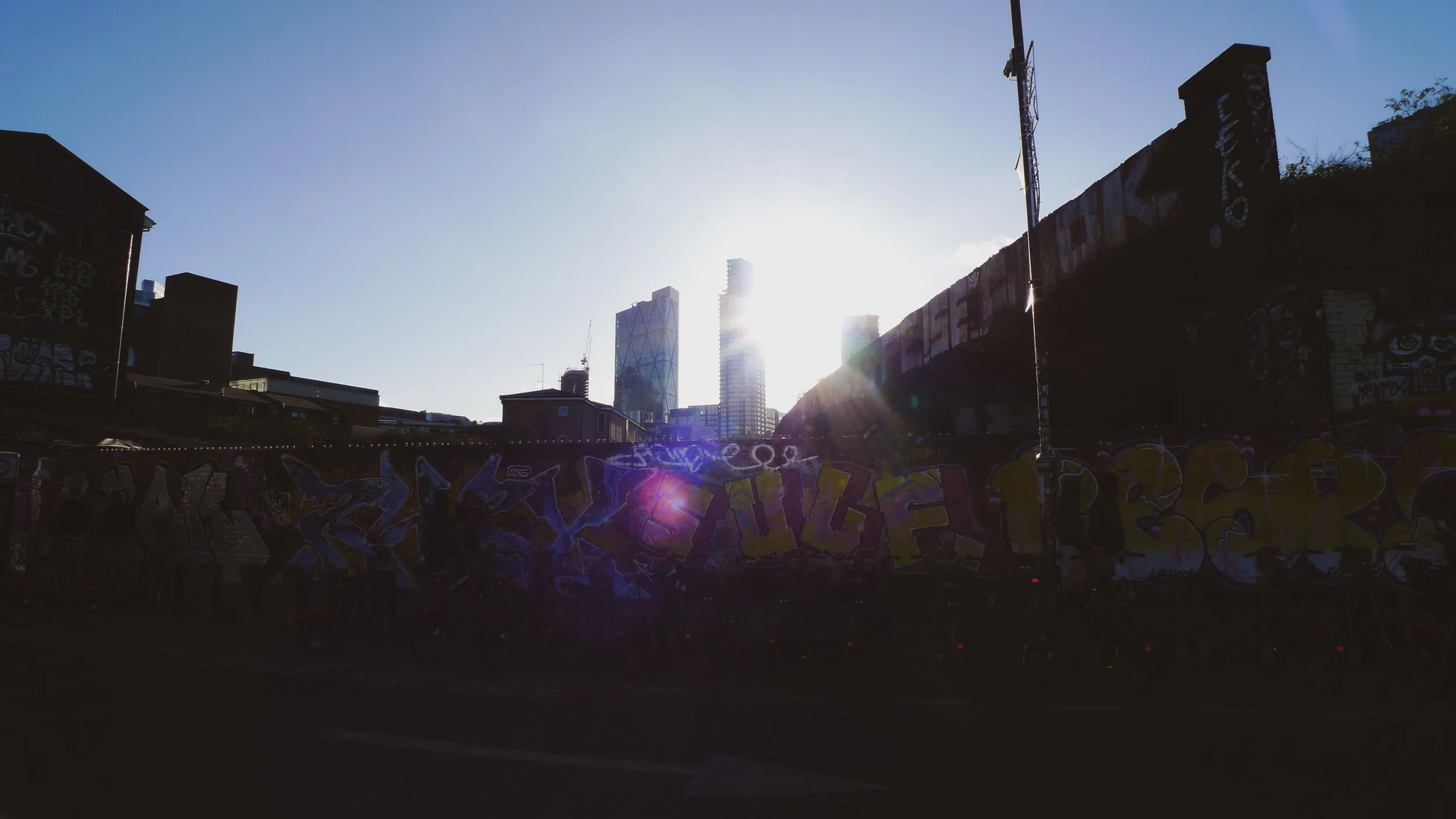 Cityscape with tall modern buildings in sunlight, graffiti-covered wall in foreground, clear blue sky.