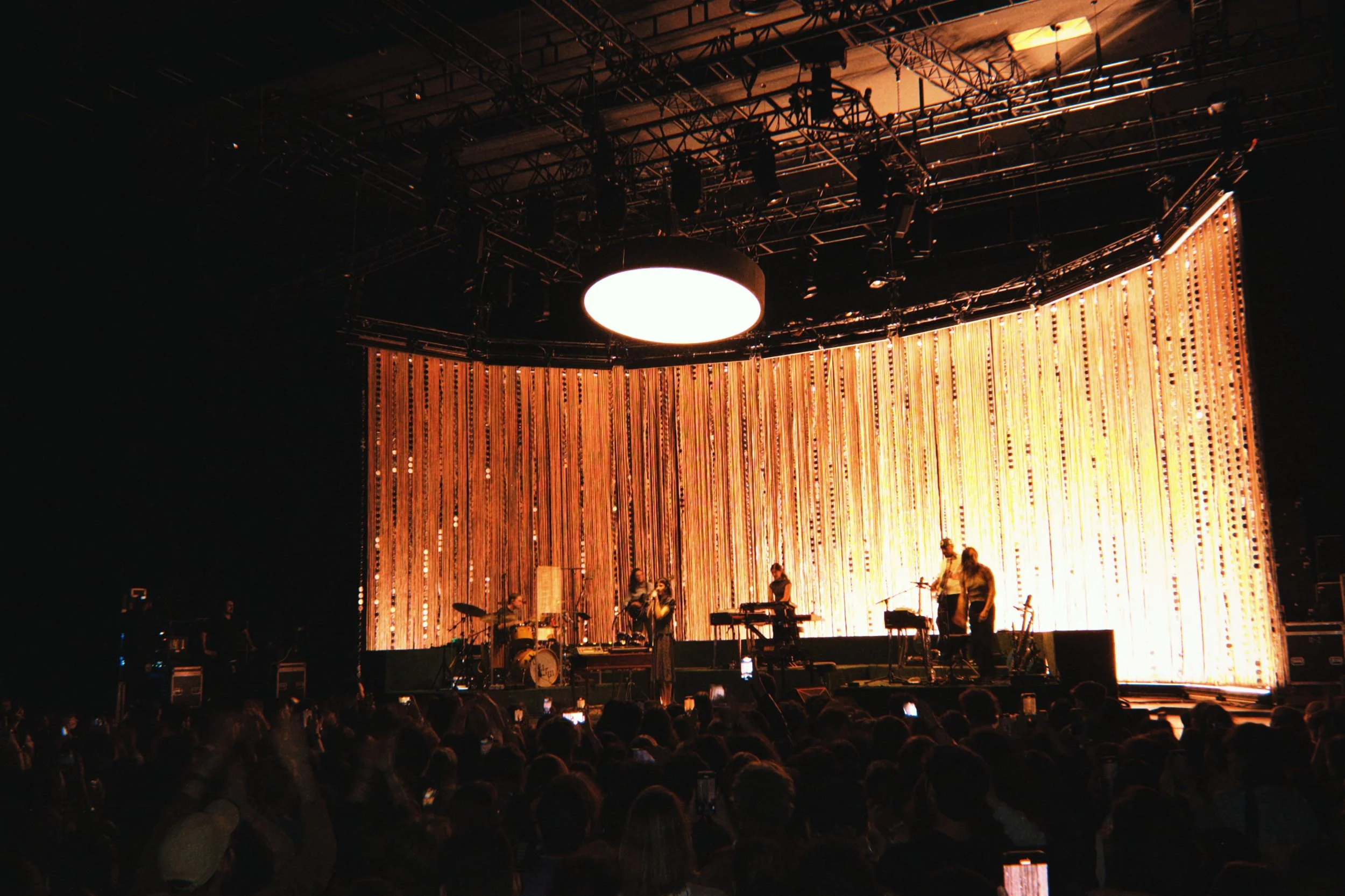 A stage set up for a concert with musical instruments and performers, backdrop with hanging orange strips, overhead lighting, and a large round light fixture.