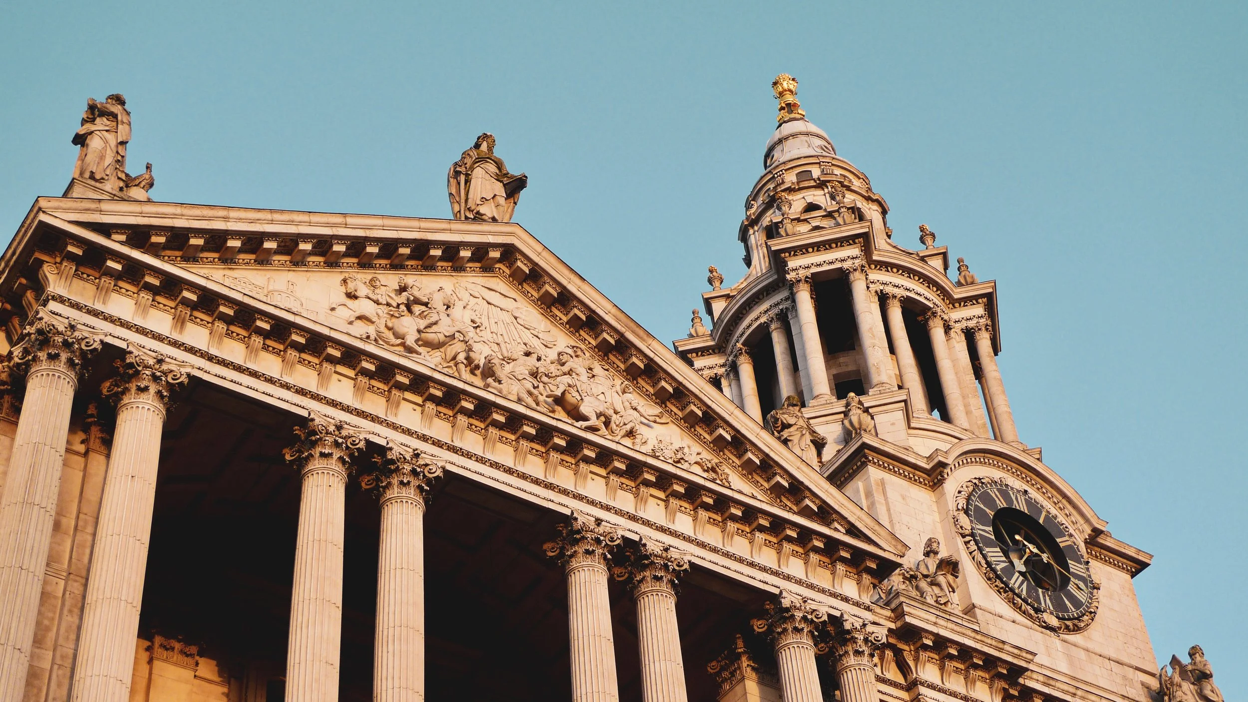 Close-up of an ornate, historic building with large columns, statues on the roof, and a clock on the tower, set against a clear blue sky.