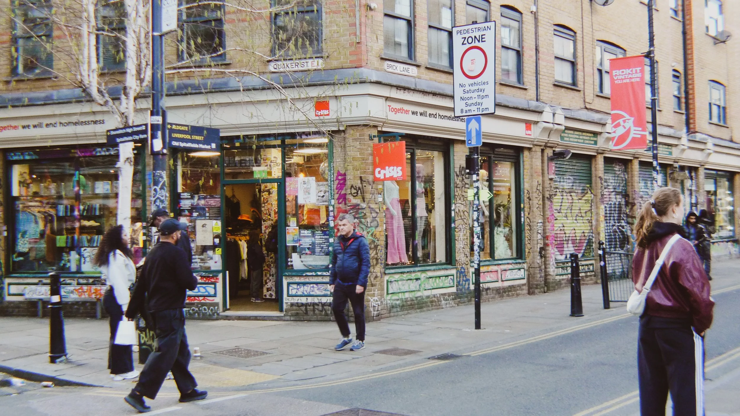 Street scene with people walking past a graffiti-covered storefront on a corner. Street signs for Quaker Street and Brick Lane are visible, with graffiti and posters on the building. Pedestrian zone sign indicates restrictions on Saturdays and Sunday