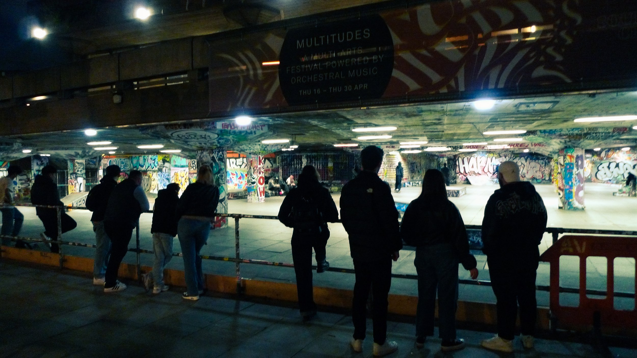 People waiting in line inside a graffiti-covered skate park, illuminated by overhead lights, with a banner advertising a multi-arts festival.