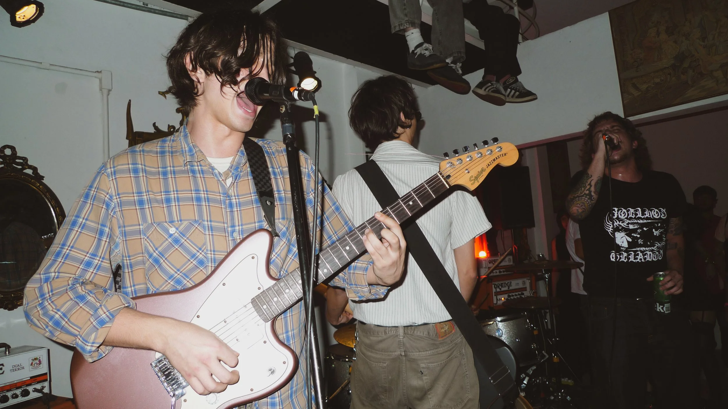 Group of young men performing live music at an indoor venue, with one playing an electric guitar and others singing into microphones.