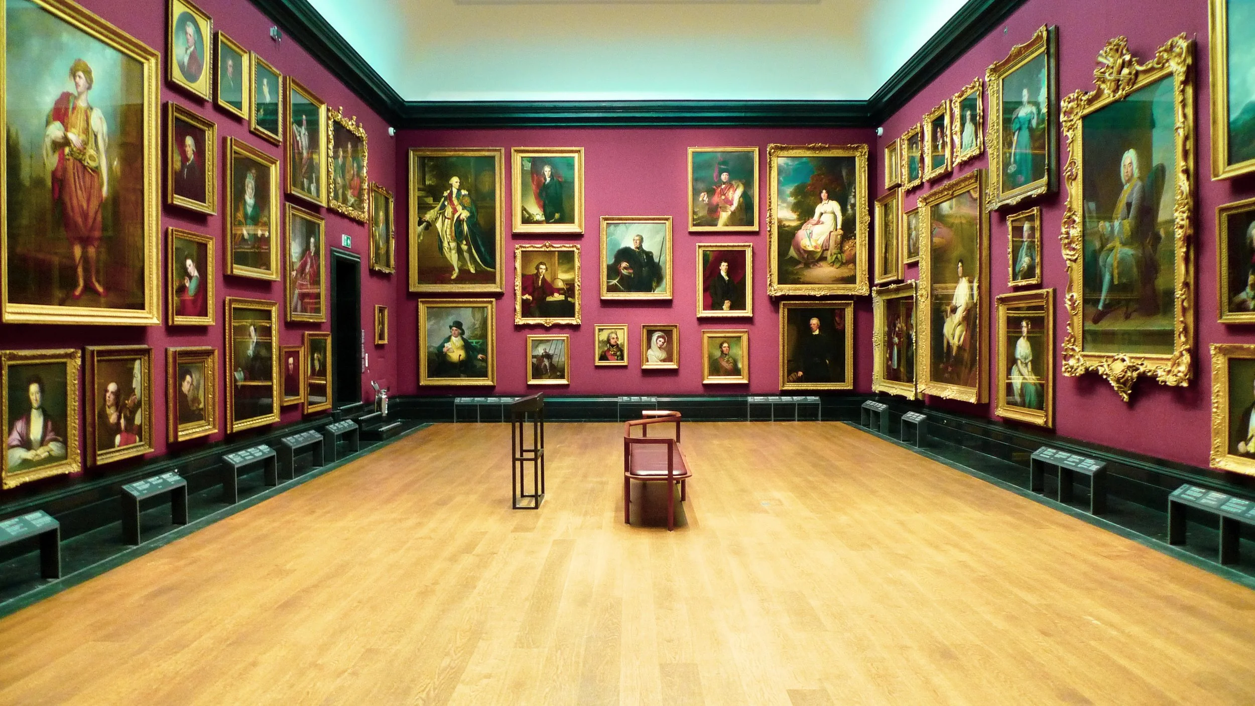 An art gallery with red walls displaying numerous framed oil portraits of historical figures. The room has a wooden floor, a few chairs, and small information plaques beneath the paintings. The ceiling is white and there is green trim along the top a