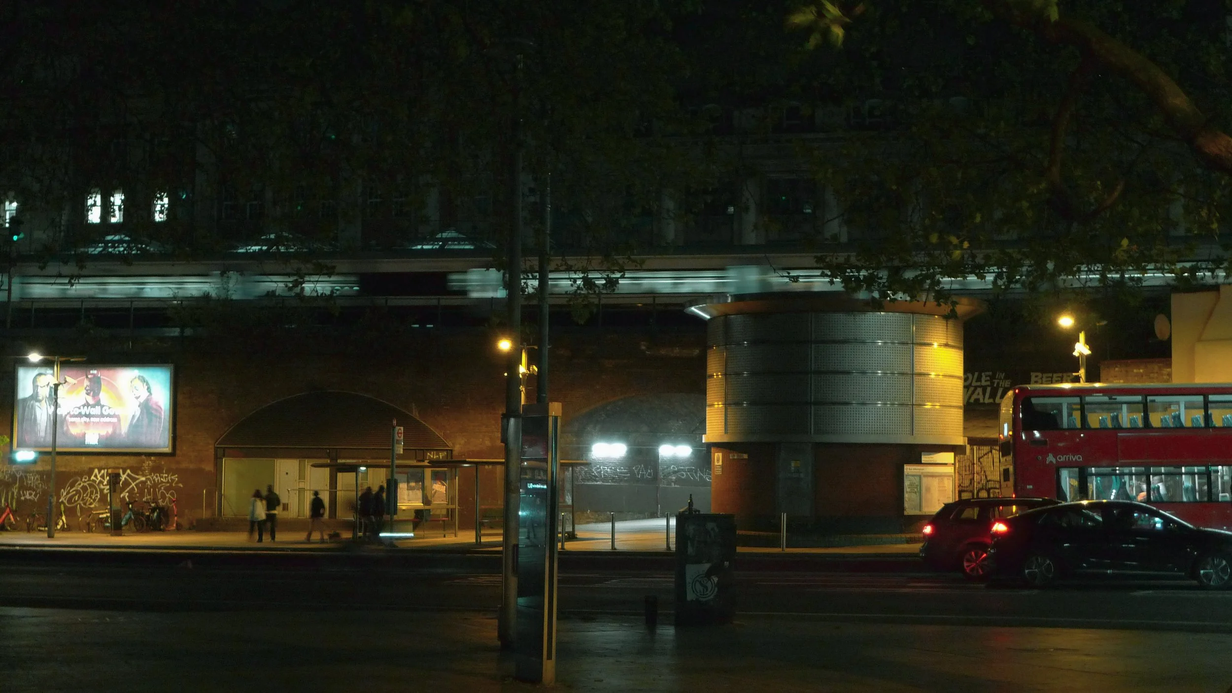 Nighttime scene of a city street with a bus stop, people walking, and a double-decker bus. A billboard and graffiti are visible on a brick building. Tree branches canopy above.