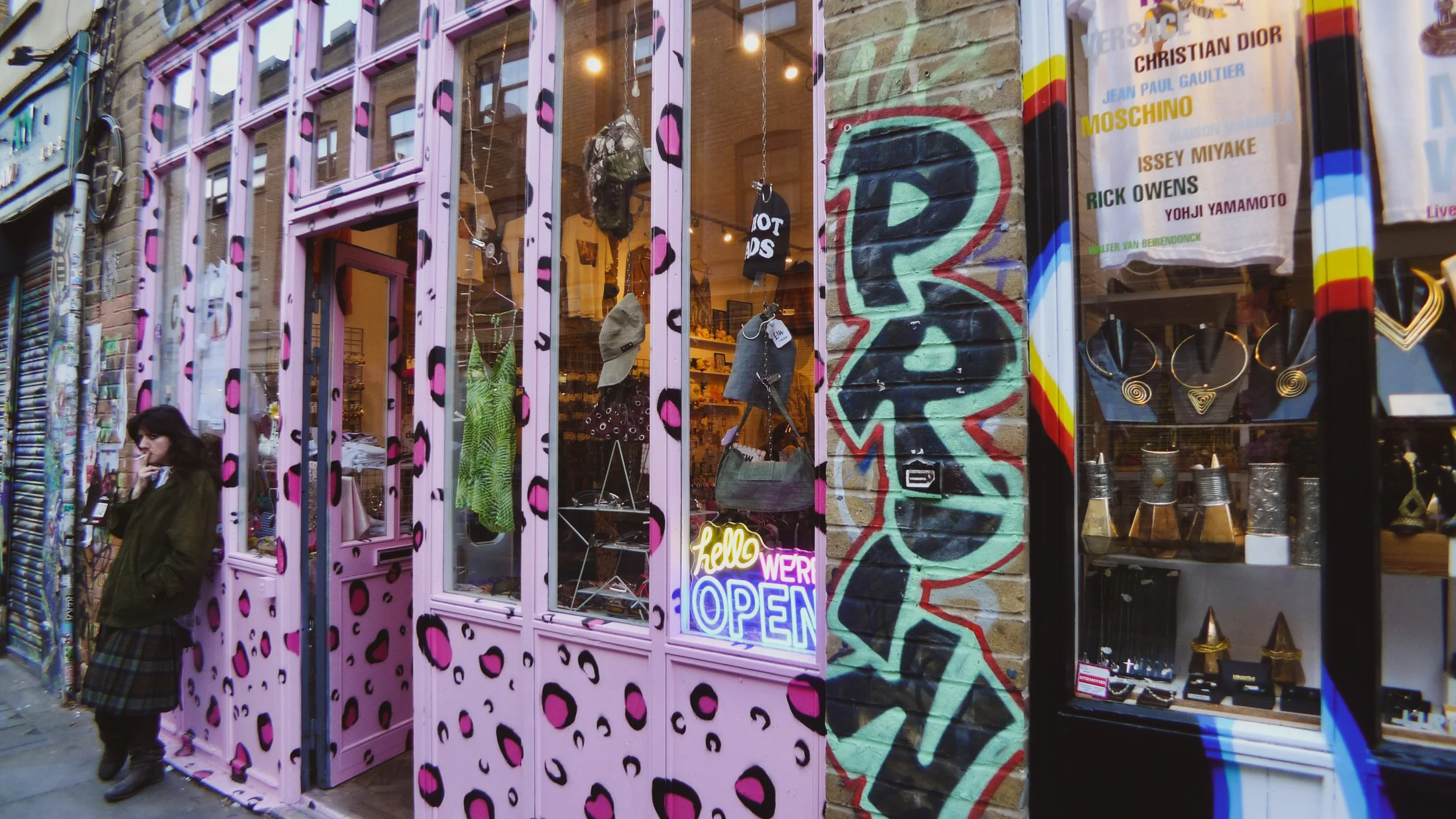 A storefront with pink and black leopard print decor and a neon sign that says 'Hello We're Open'. The window displays various jewelry, clothing, and accessories. A woman is standing outside the shop, looking at her phone.