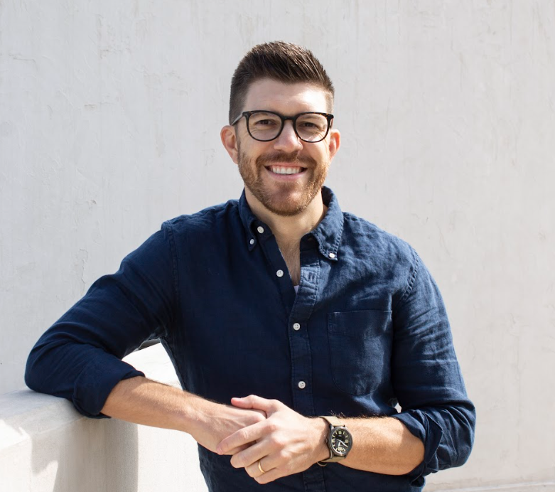 A smiling man with glasses, a beard, wearing a navy blue button-up shirt, leaning on a white surface against a light-colored wall.