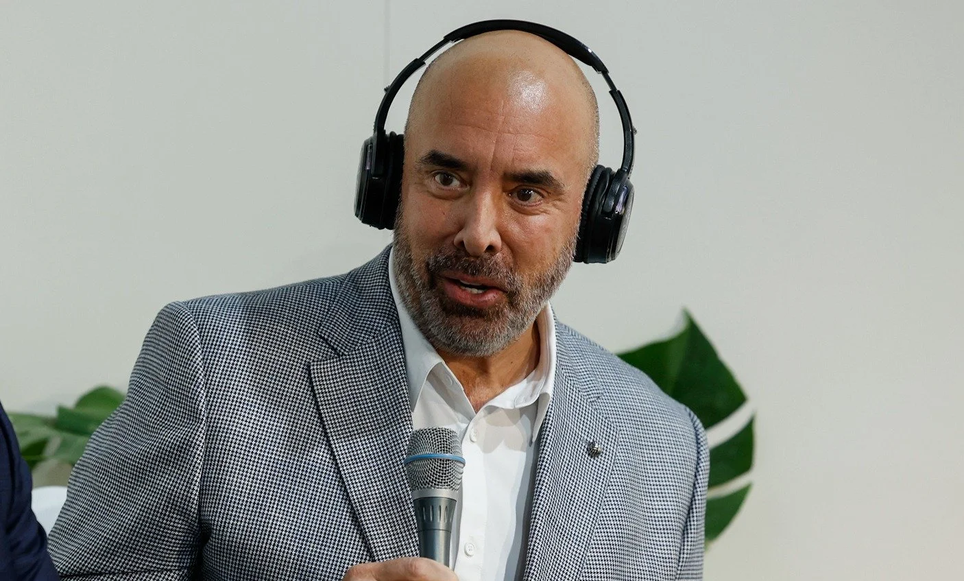A middle-aged man with a gray beard and bald head, wearing a checkered blazer and white shirt, holding a microphone and wearing large black headphones, speaking at an indoor event.