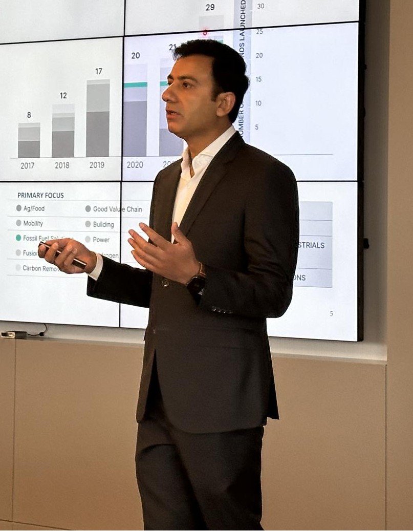 Man in a dark suit giving a presentation with a large screen behind him showing bar graphs and data charts.