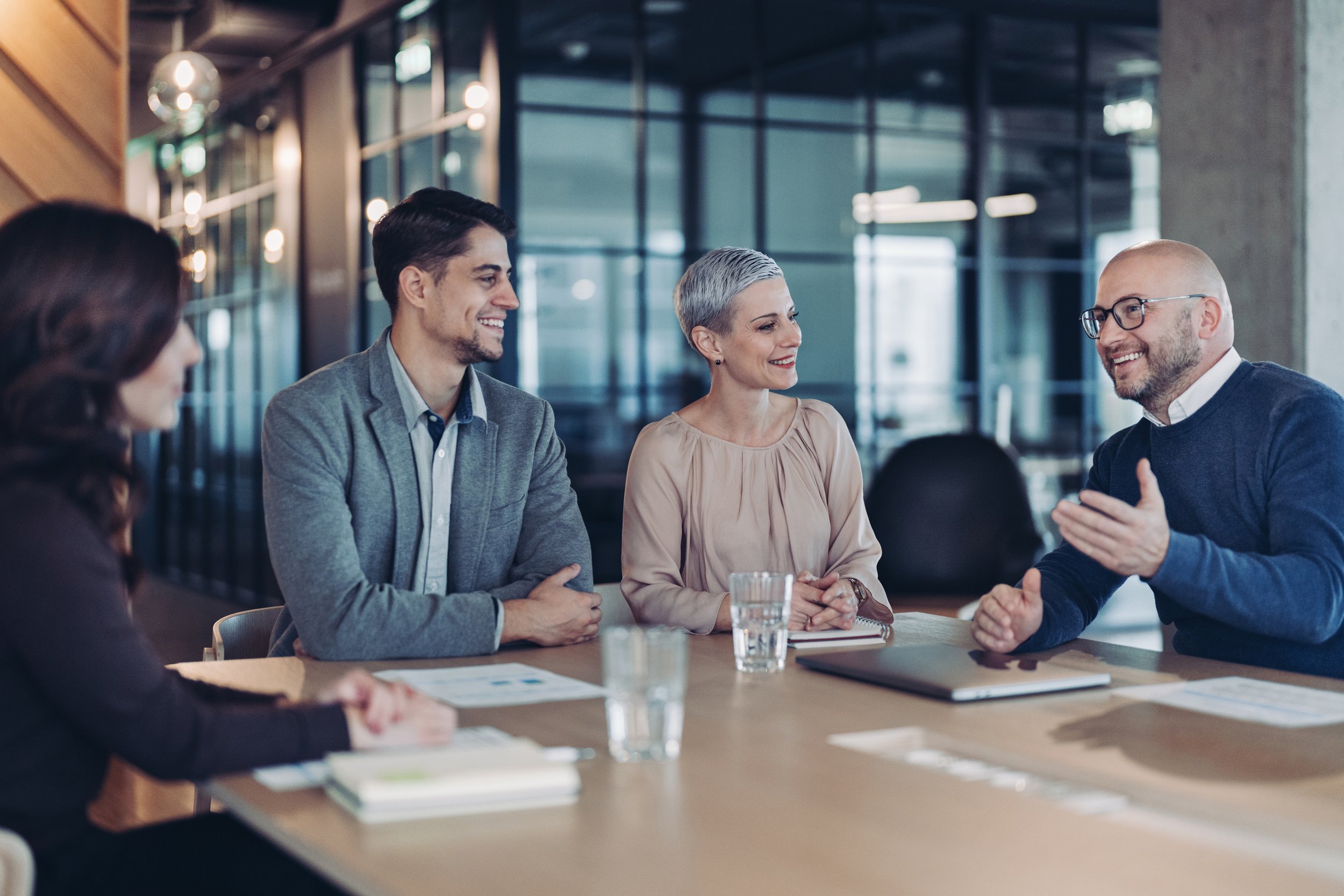 Group of five professionals having a business meeting in a modern conference room with glass walls, smiling and engaging in conversation.