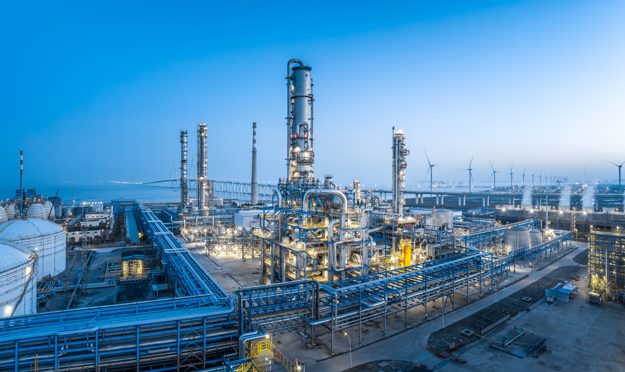 Oil refinery with multiple towers, pipelines, and wind turbines in background during dusk.