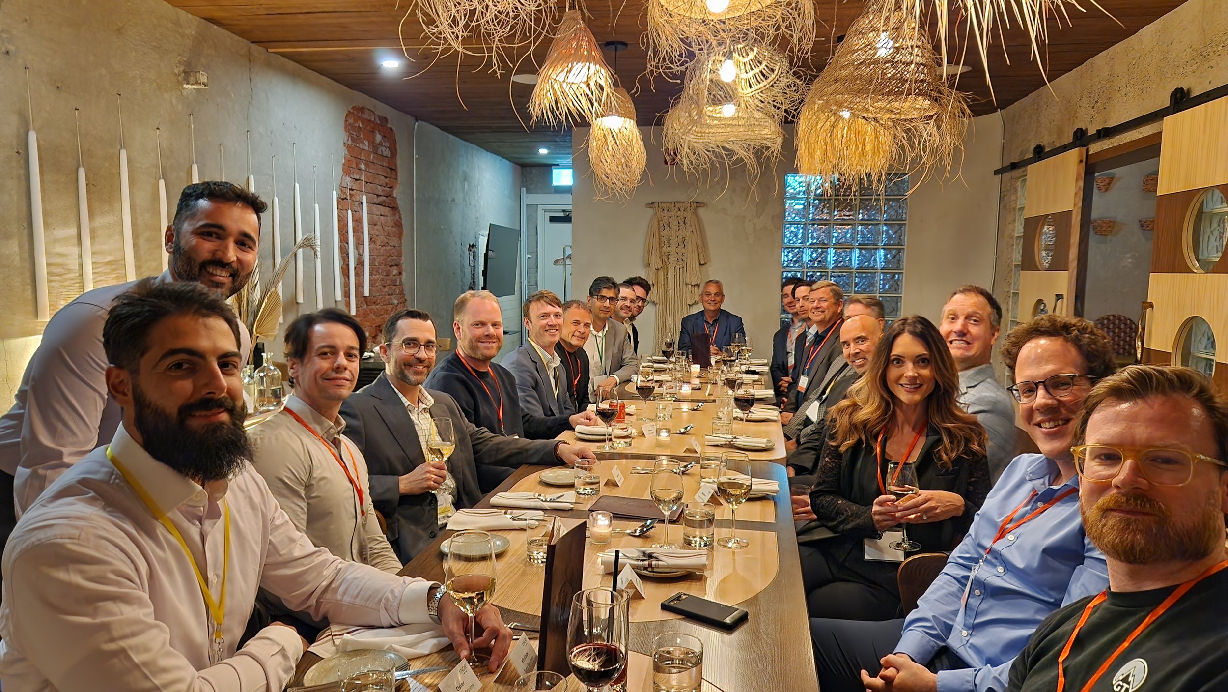 Group of people sitting around a long dining table, smiling, at a formal gathering or event, with drinks and table settings, in a warmly lit restaurant with decorative hanging lamps and rustic decor.