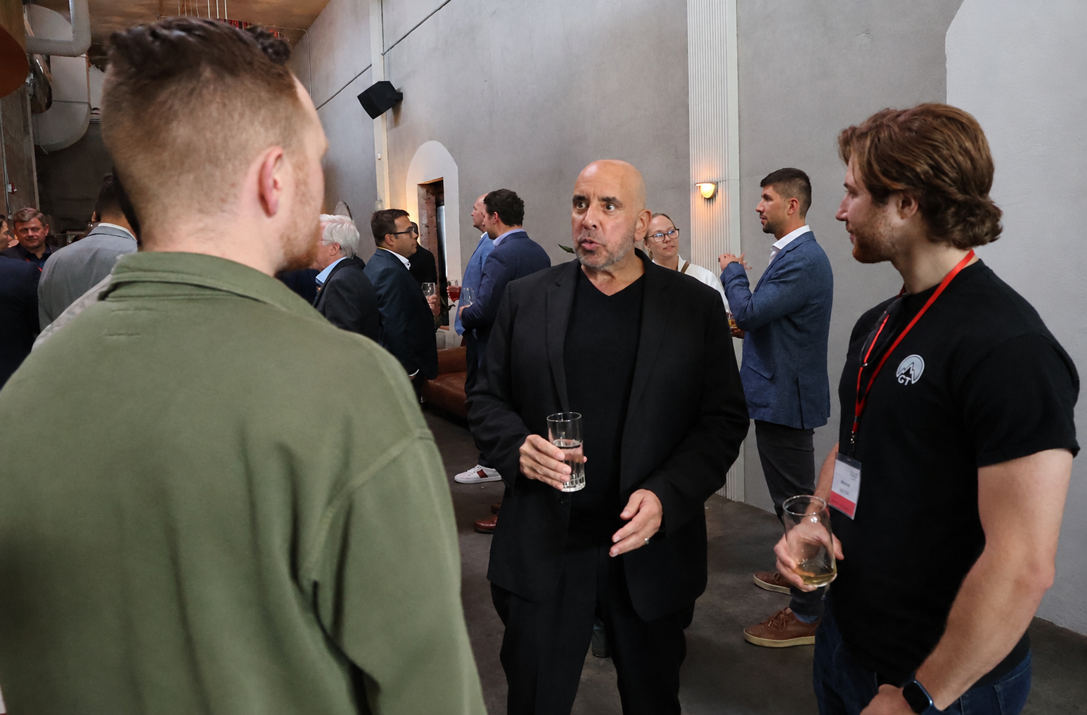 A group of men and women at a social event, engaged in conversation, standing in a room with gray walls and warm lighting, holding drinks.