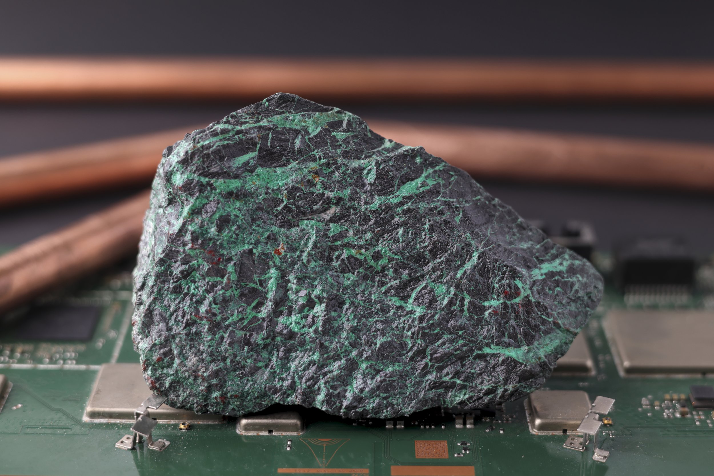 A large raw mineral with black and green marbled patterns sits on a circuit board with electronic components and copper wires in the background.