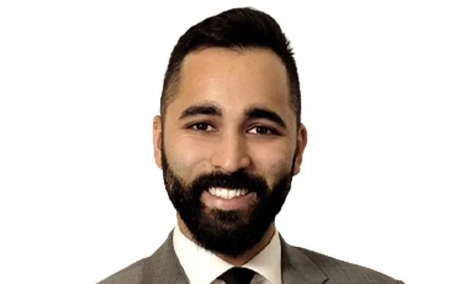 A smiling man with dark hair and a beard, dressed in a gray suit and white shirt, against a plain white background.