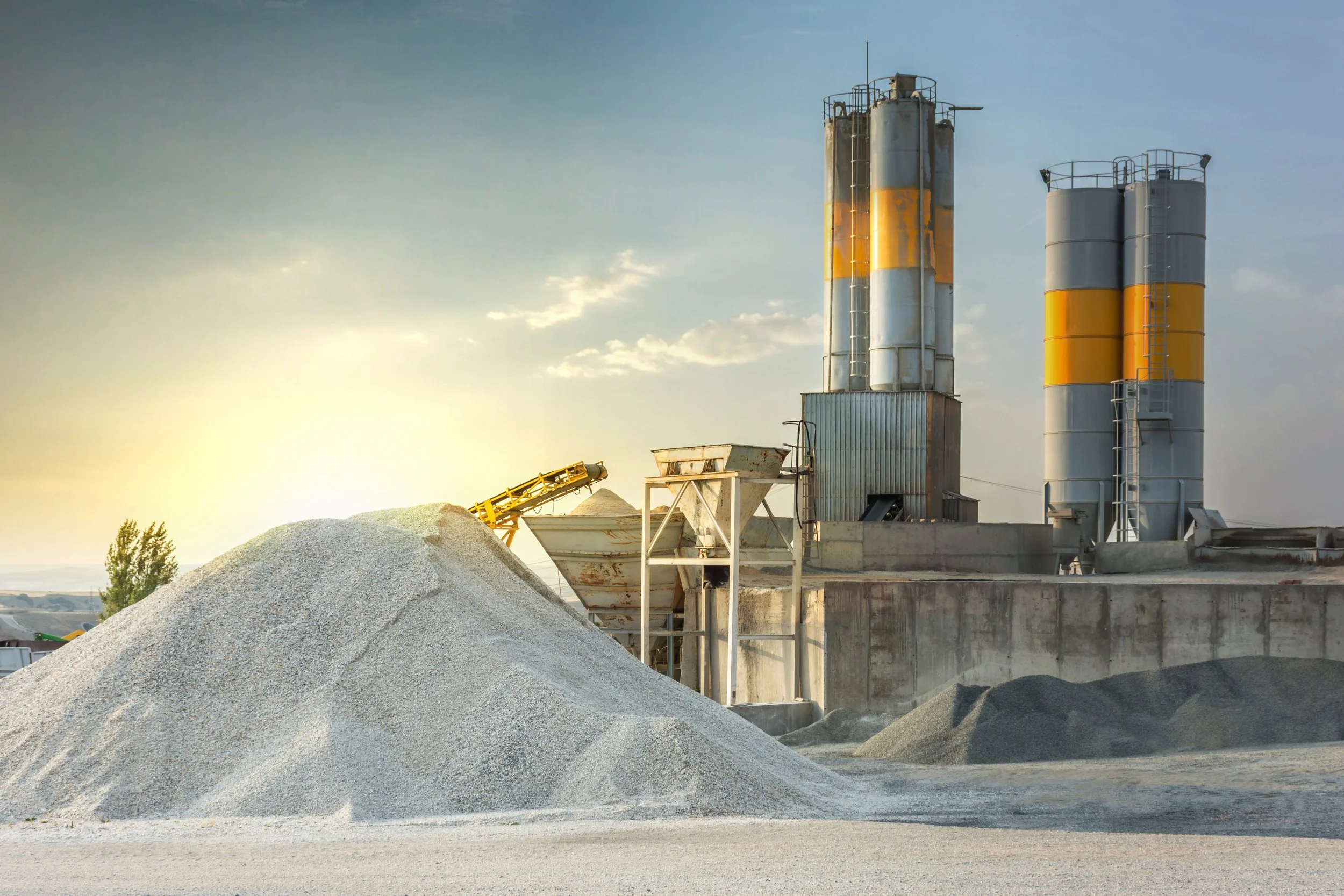 Industrial site with large gravel pile, metal machinery, and smokestacks with yellow and gray stripes, under a clear sky with sunlight.