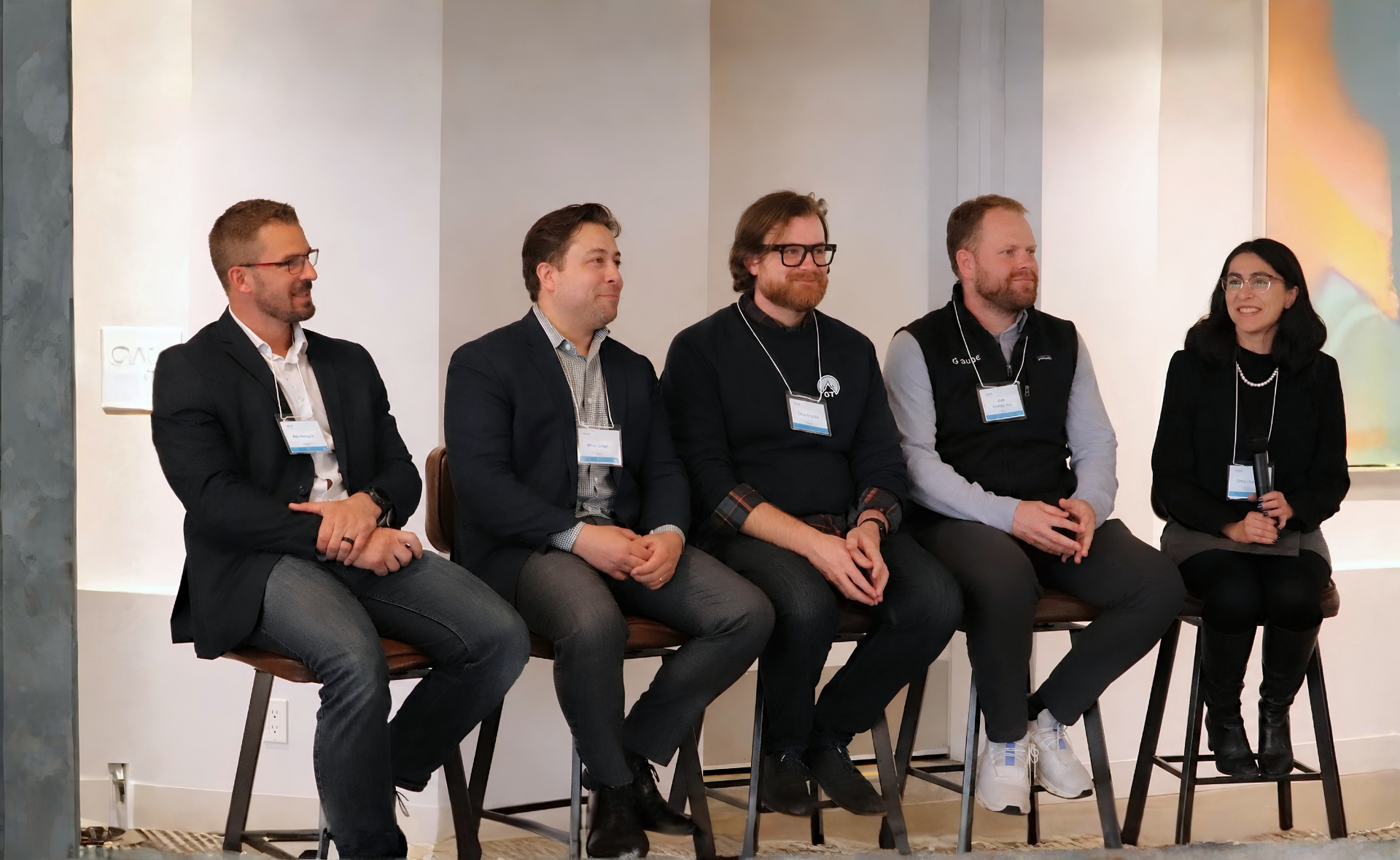 Five people sitting on barstools during a panel discussion at a conference, with three men and two women, all wearing business casual attire and name tags.