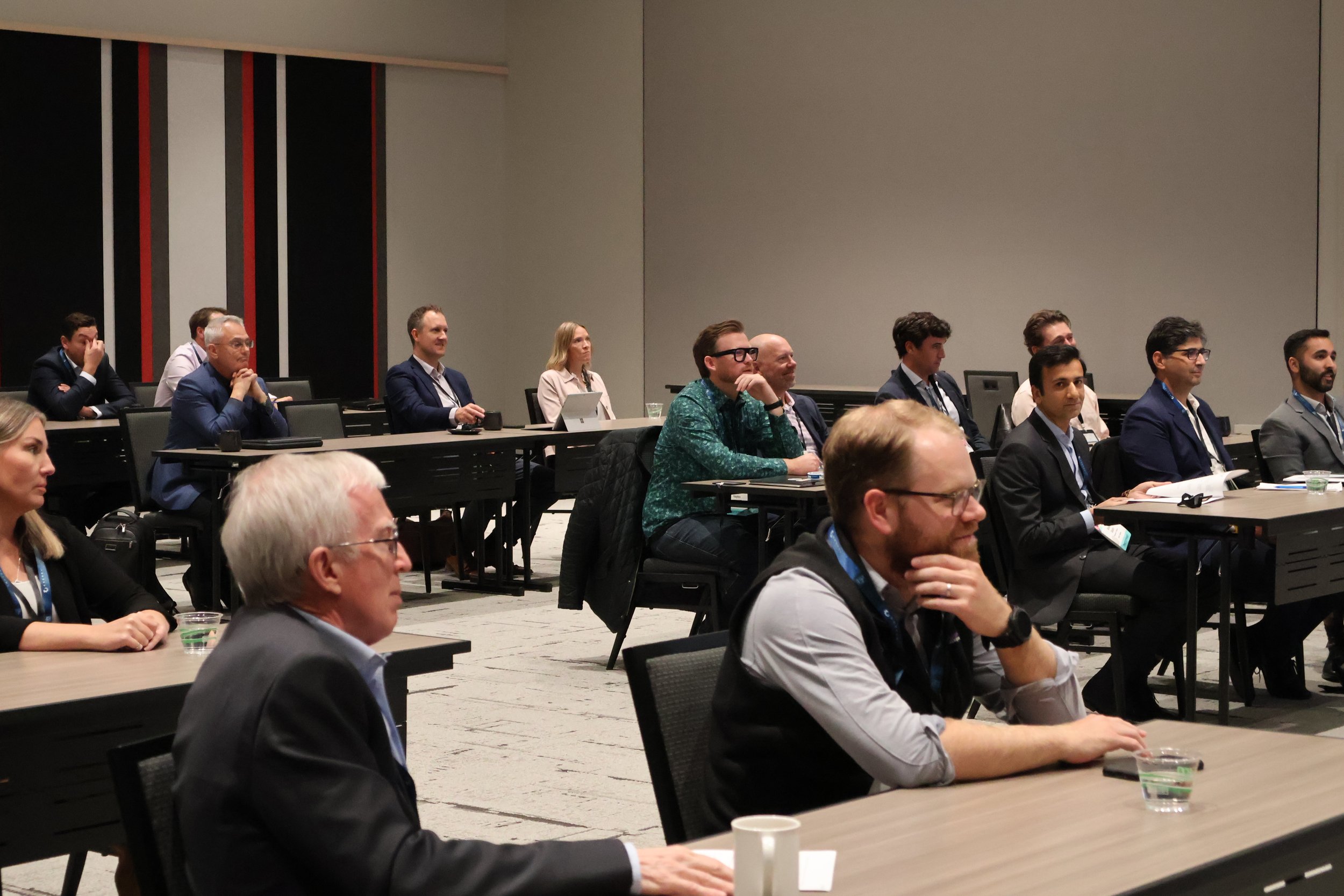 A group of diverse professionals attending a conference, seated at tables in a large modern conference room, attentively listening to a speaker or presentation.
