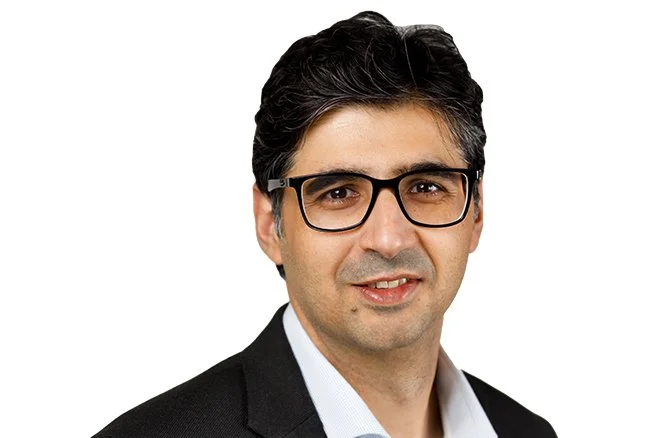 Portrait of a man wearing glasses and a dark suit, smiling against a white background.