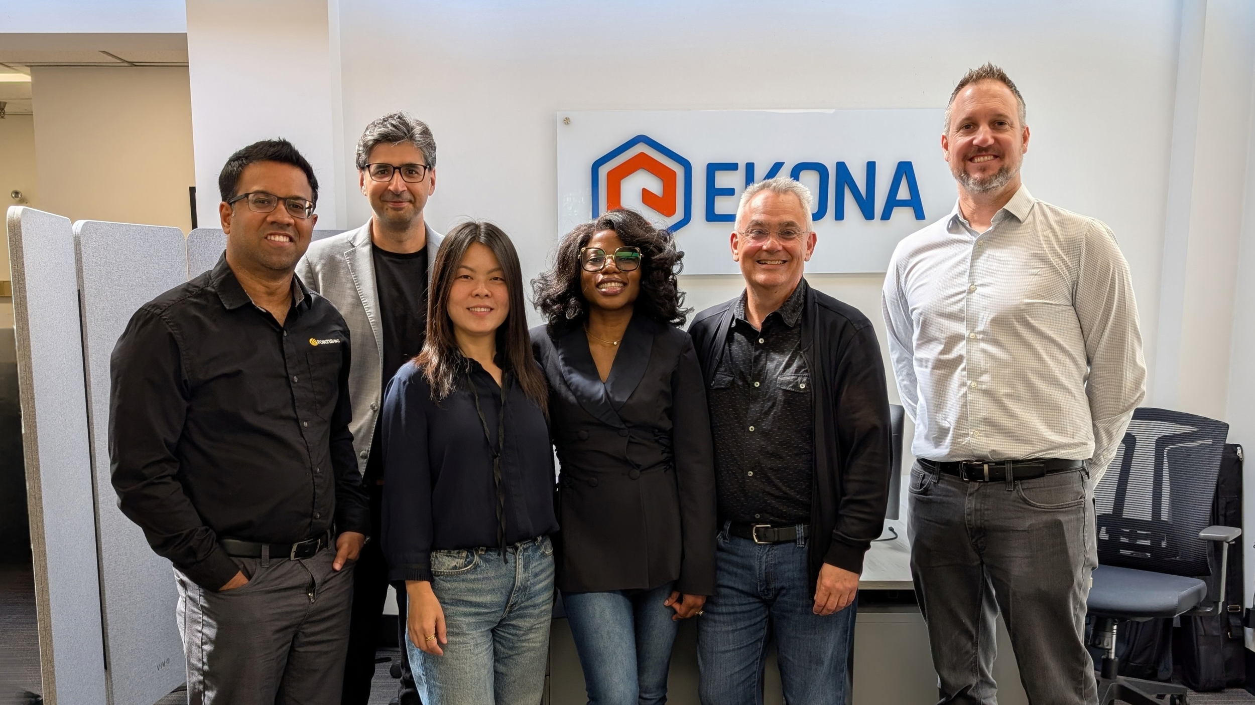 Group of six diverse professionals standing together in front of a sign that reads 'EKONA' at an office or conference room.