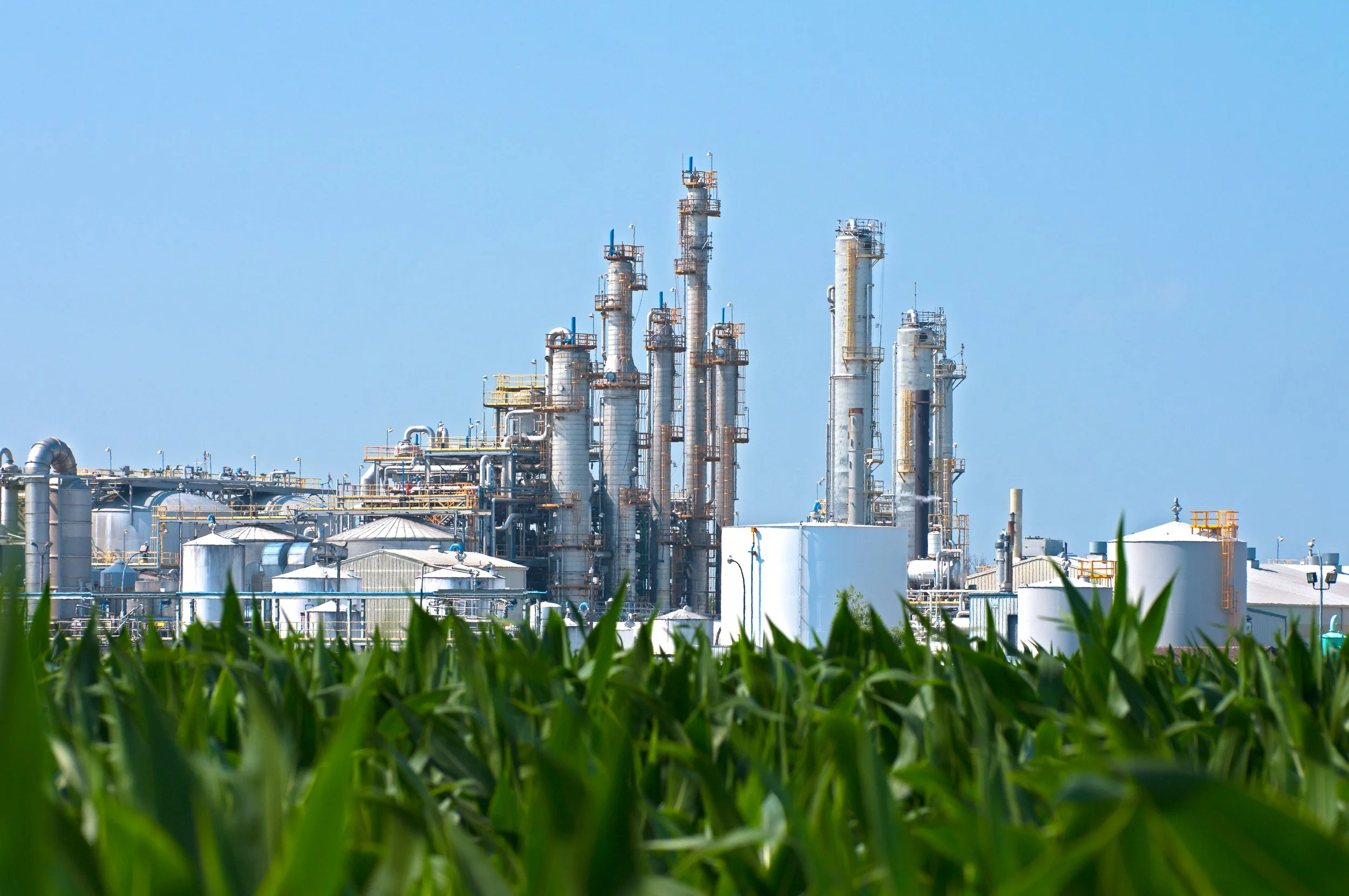 An industrial factory with tall white smokestacks and metal tanks viewed above a green cornfield against a clear blue sky.