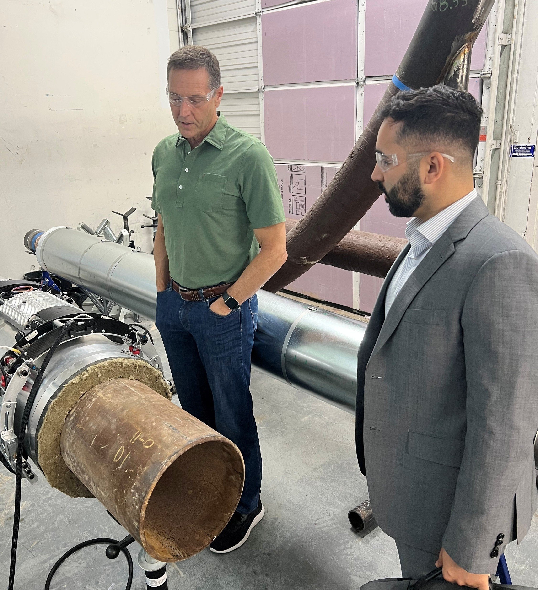Two men in safety glasses examining a large pipe with mechanical and electronic equipment in an industrial setting.
