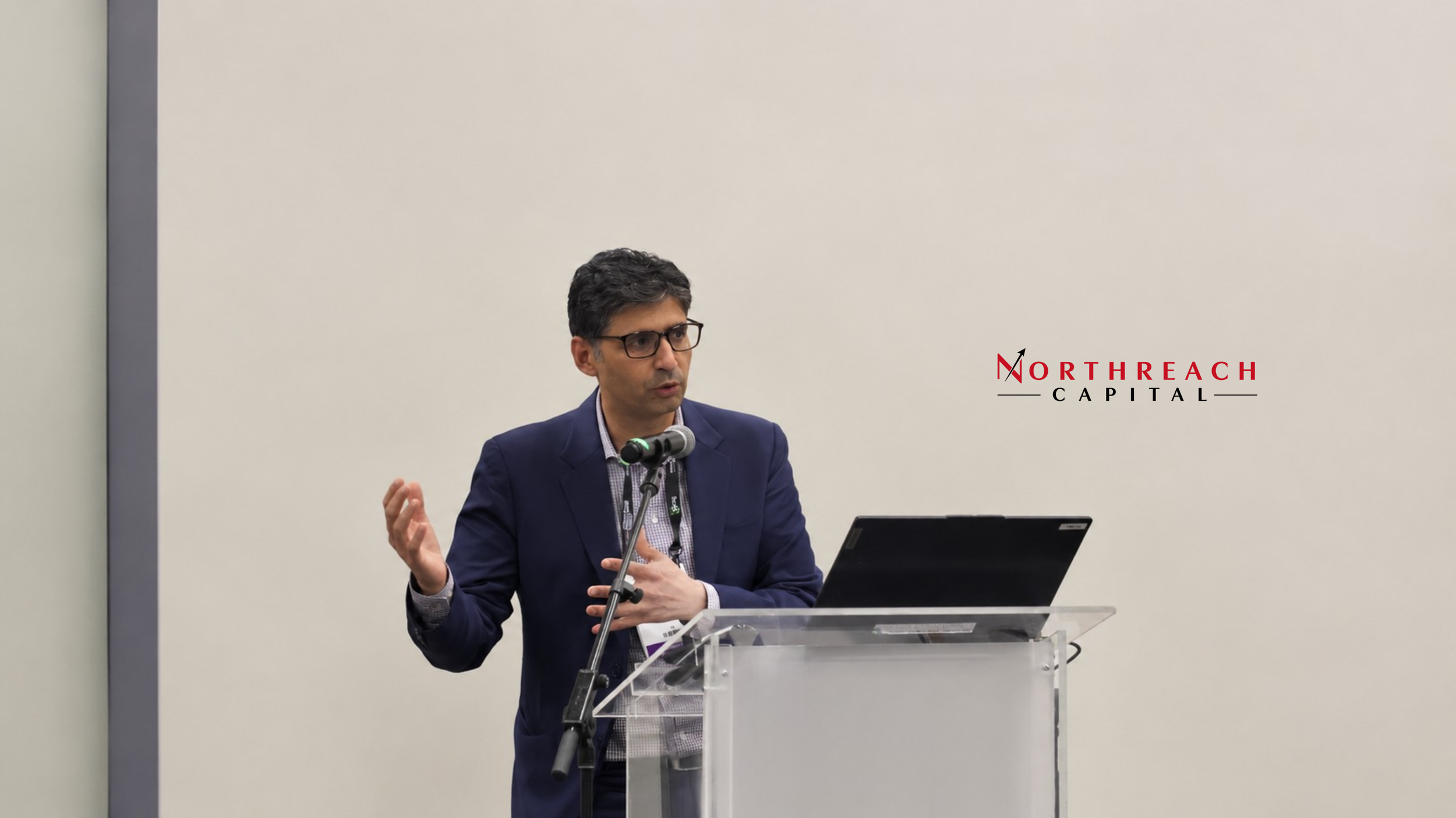 A man in a navy blazer and glasses speaking at a podium with a microphone, in front of a white wall with the red and black logo of 'Northreach Capital'.