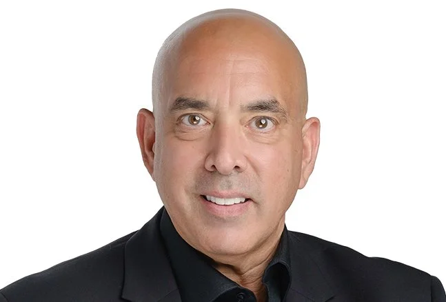 A bald man wearing a black suit and black shirt, smiling against a white background.