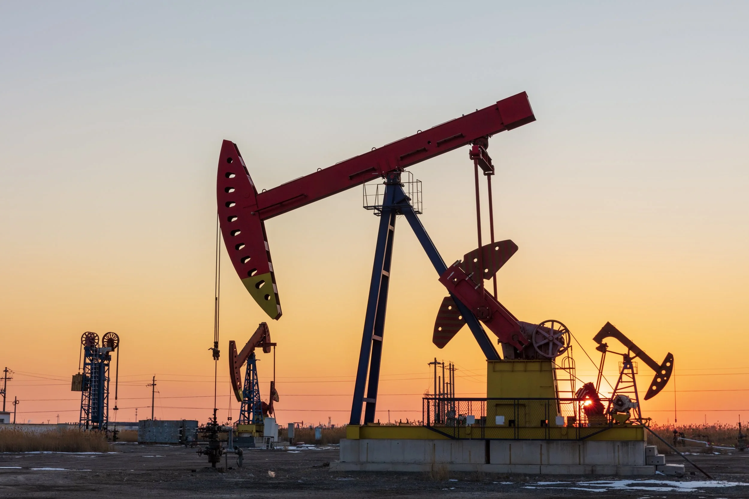 Multiple oil pumpjacks operating as the sun sets or rises in the background at an oilfield.