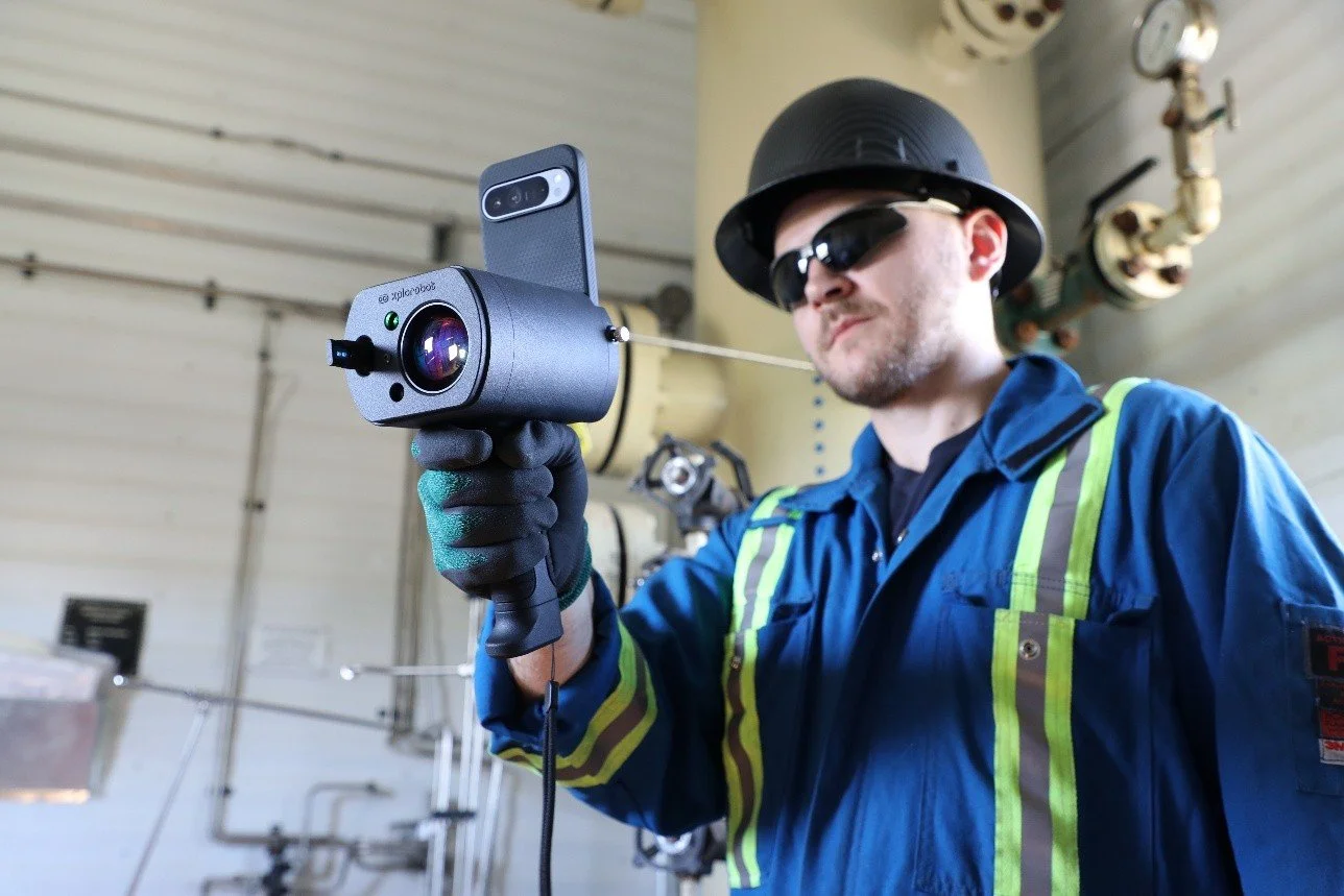 A man wearing safety glasses, a helmet, and a blue work jacket holding a camera with a smartphone attached, aimed at an industrial setting with pipes in the background.