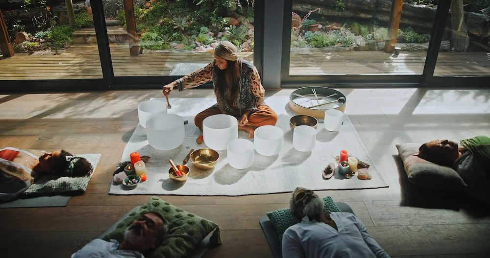 A woman is sitting on a white rug playing crystal singing bowls, surrounded by candles and bowls, with four people lying on cushions around her. Schedule a Sound Bath session with Synergy Healing Arts in Ogden. Utah.