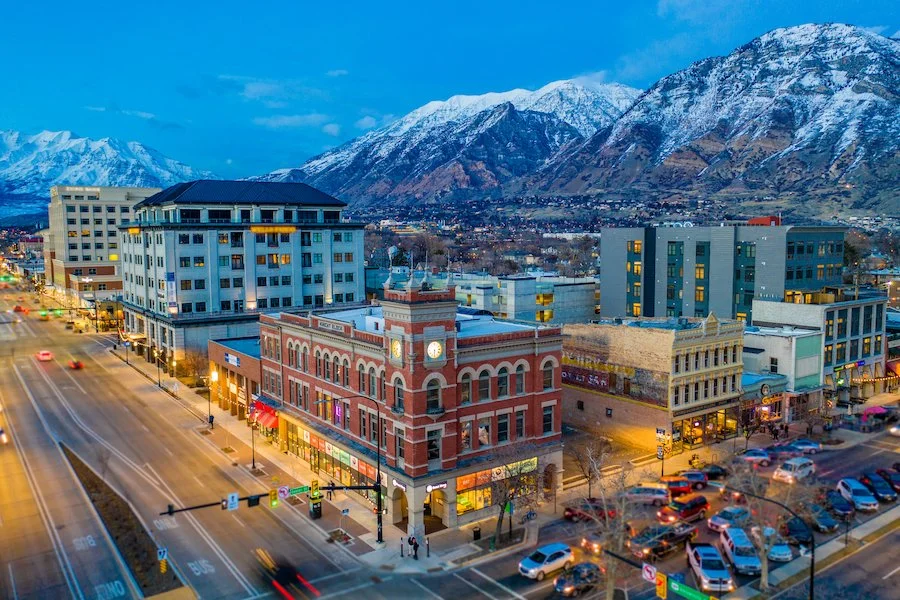 Downtown street with parked cars, commercial buildings, and snow-capped mountains in the background during evening. Book a Mobile Sound Bath with Synergy Healing Arts in Ogden, Utah.