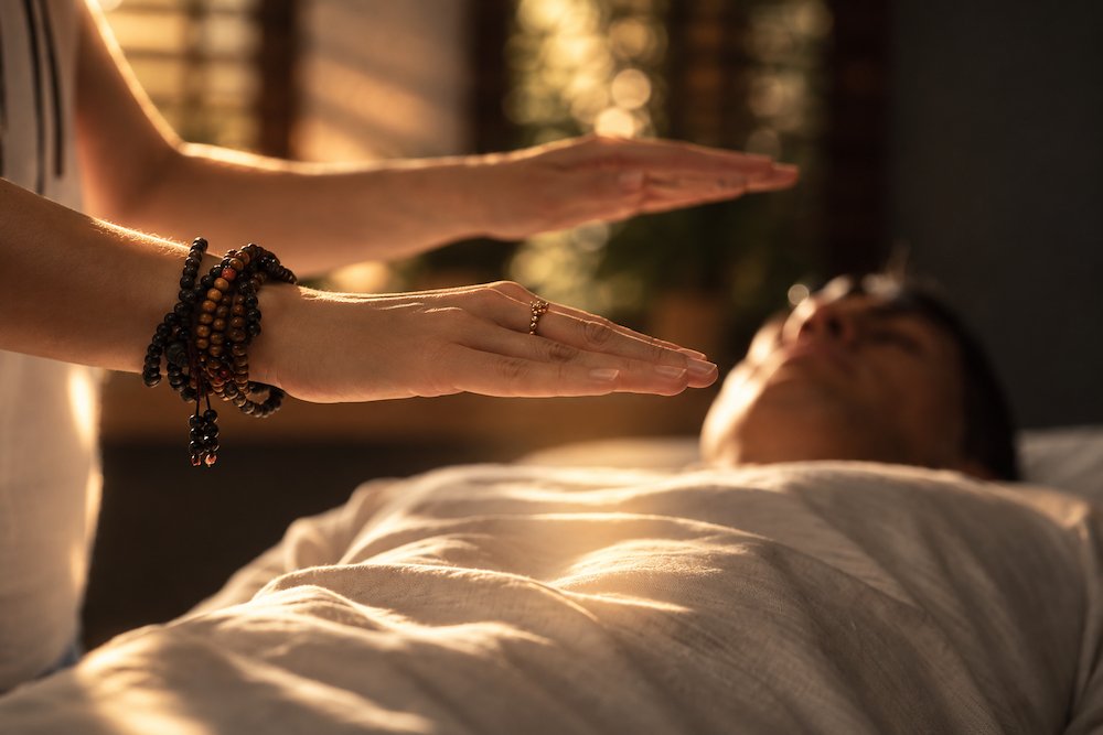 Person performing a healing or energy healing (reiki) ritual on another person lying in bed, with hands raised above the person. Schedule a Reiki Healing session with Synergy Healing Arts in Ogden. Utah.