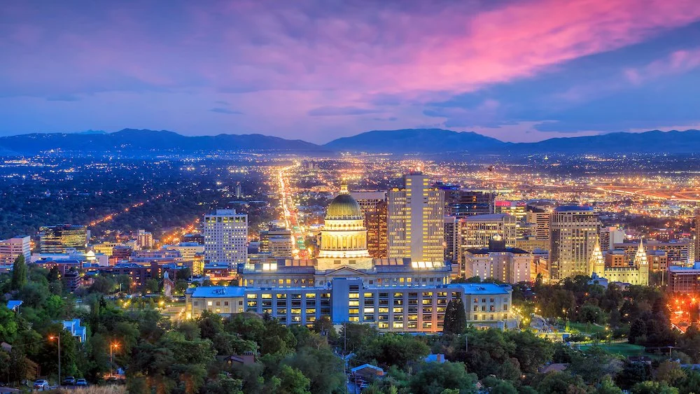 Night cityscape of Denver, Colorado, with illuminated buildings and mountains in the background, under a purple and pink sky. Book a Mobile Sound Bath with Synergy Healing Arts in Ogden, Utah.