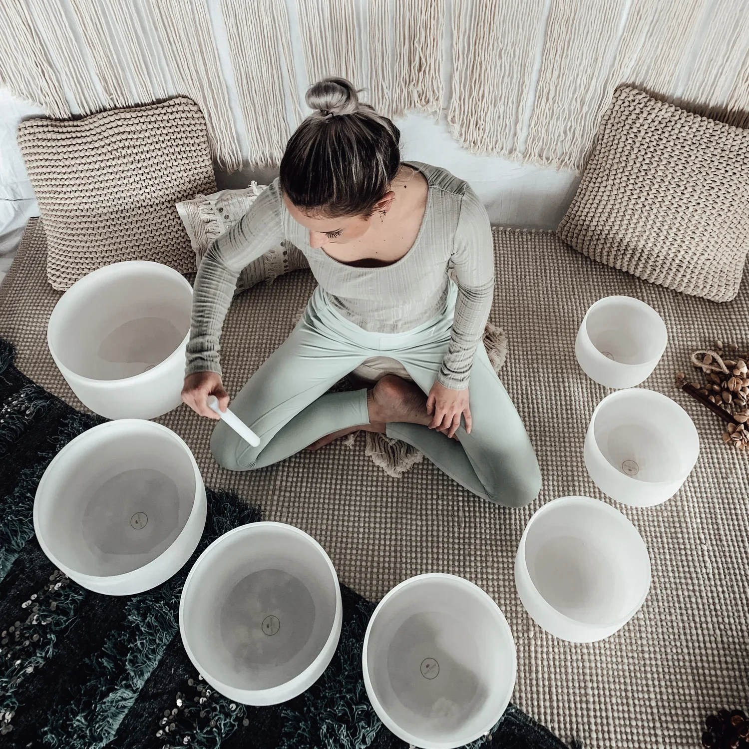 A woman sitting on a beige textured rug surrounded by empty singing bowls and decor pillows, preparing to play the bowls with a mallet.  Book a Sound Bath at Synergy Healing Arts in Ogden, Utah.
