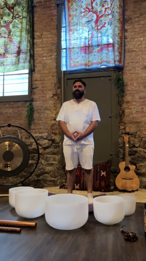 A man with a beard dressed in white standing in front of a brick wall, with singing bowls in front, a gong to his left, an acoustic guitar, colorful fabric window coverings, and decorative textiles in an industrial-style space.