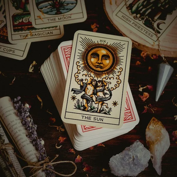 A tarot card deck with the Sun card on top, surrounded by other tarot cards, crystals, dried flowers, and a small metallic cone on a dark wooden surface.  Book a Tarot Reading at Synergy Healing Arts in Ogden, Utah.