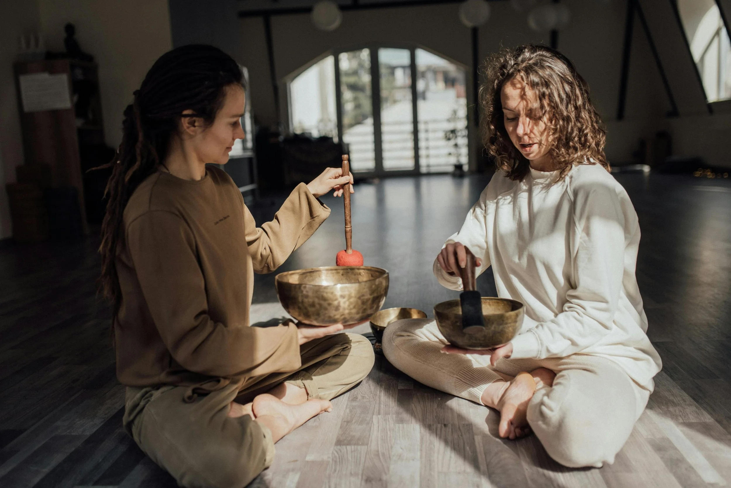 Two women sitting cross-legged on the floor, playing singing bowls in a warmly lit room with large windows. Schedule a Sound Bath session with Synergy Healing Arts in Ogden. Utah.