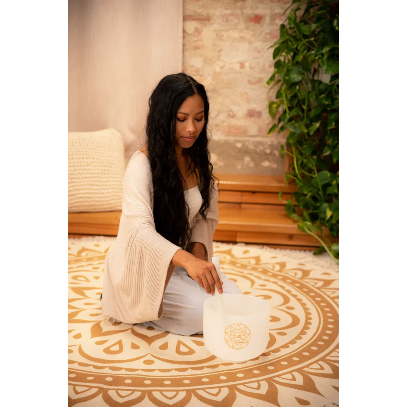 A woman with long black hair sitting cross-legged on a decorative patterned rug in a cozy room, playing a crystal singing bowl decorated with a geometric design. Book a Sound Bath at Synergy Healing Arts in Ogden, Utah.