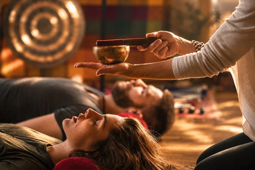 People practicing sound healing with singing bowls, lying on yoga mats in a dimly lit room. Schedule a Sound Healing session at Synergy Healing Arts in Ogden. Utah.