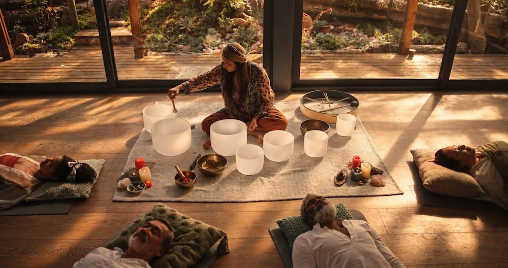 A group of five people lying on yoga mats in a sunlit room with large windows, watching a woman play crystal singing bowls on a rug with candles and bowls around her. Book a Sound Bath with Synergy Healing Arts in Ogden, Utah.